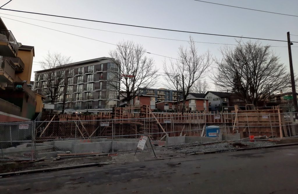 A construction pit on a triangular plot with the new Inspire Fremont building in the background across Bridge Way.