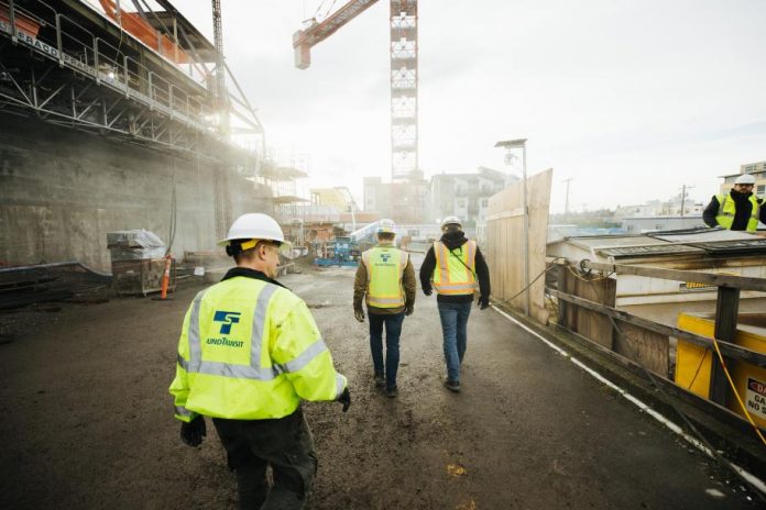 Three construction workers in Sound Transit vests walk a construction site with crane overhead.