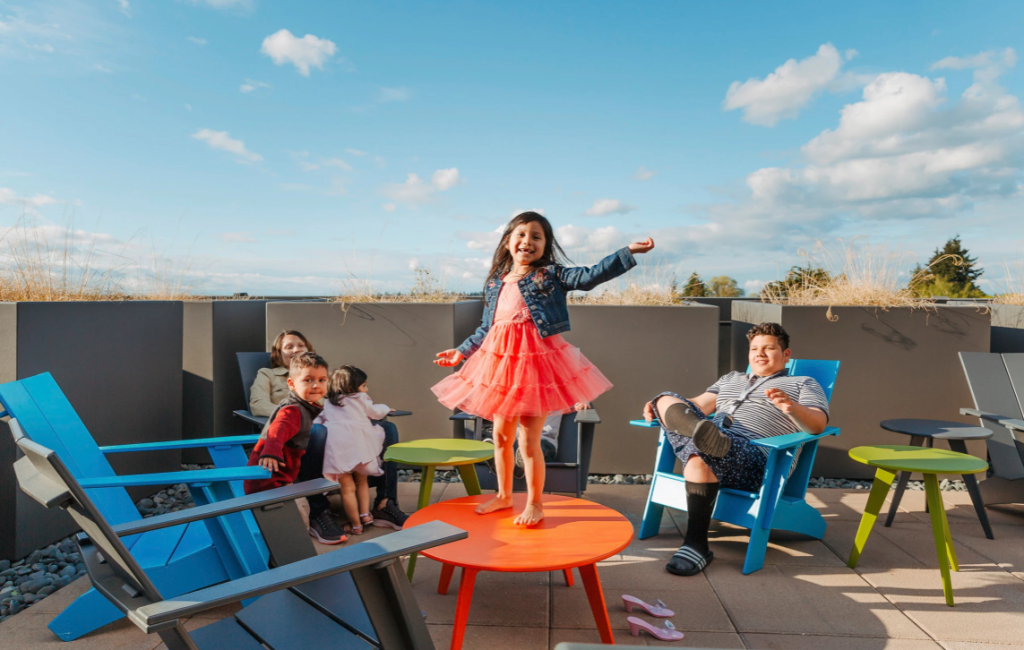 A latino family sits on patio furniture save for a girl in a tutu stands on a table.