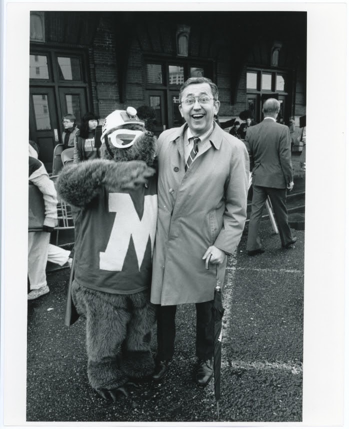 A mole mascot with goggles, a construction hat, and a big M on its chest poses with a dapper-looking man on the street.