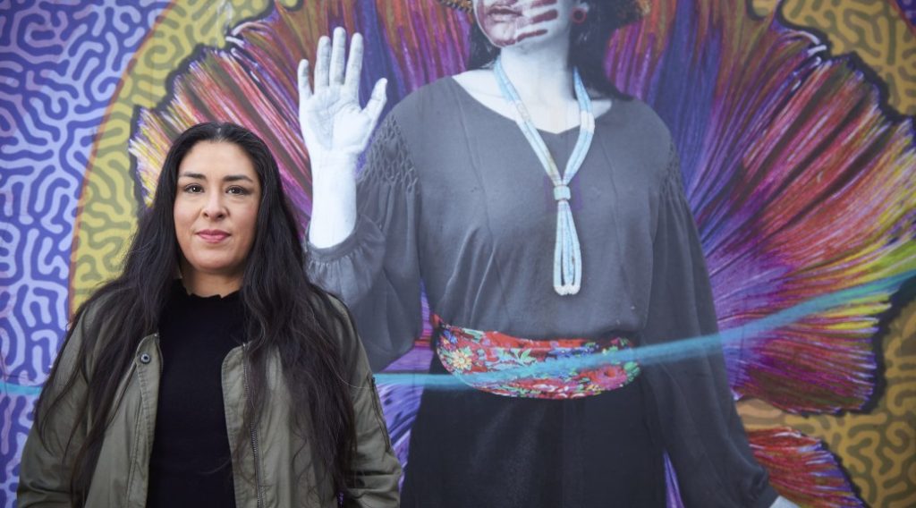 Colleen Echohawk with a colorful mural in the background showing an indigenous with a handprint over her lips.