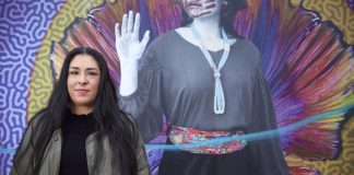 Colleen Echohawk with a colorful mural in the background showing an indigenous with a handprint over her lips.