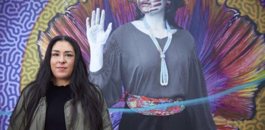 Colleen Echohawk with a colorful mural in the background showing an indigenous with a handprint over her lips.