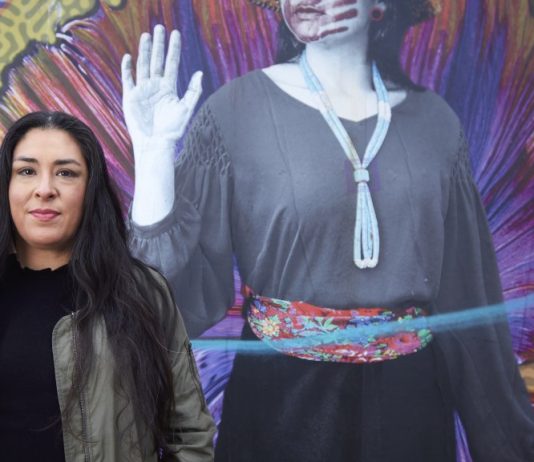 Colleen Echohawk Focuses on Housing the Homeless in Seattle Mayoral Run Colleen Echohawk with a colorful mural in the background showing an indigenous with a handprint over her lips.