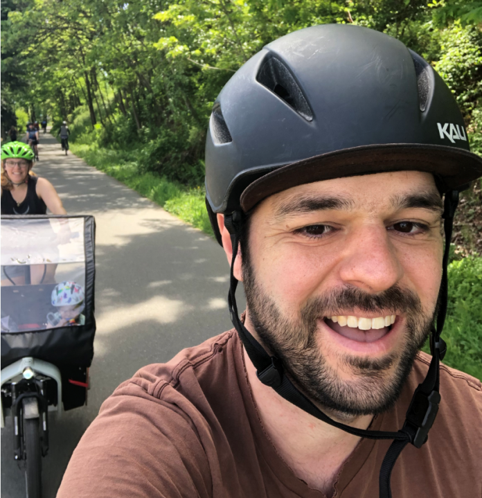 Tom taking a selfie biking on a trail with Kelli and the kid in tow