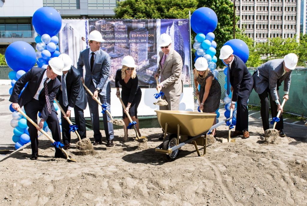 Developers in suits shovel dirt into a wheelbarrow next to a sign showing the future Lincoln Square towers.