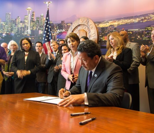 Centrists Turn to Old Formula: Crime Fears and More Money for Cops Bruce Harrell sits at a desk signing some orders at City Hall with a dozen supporters looking on.