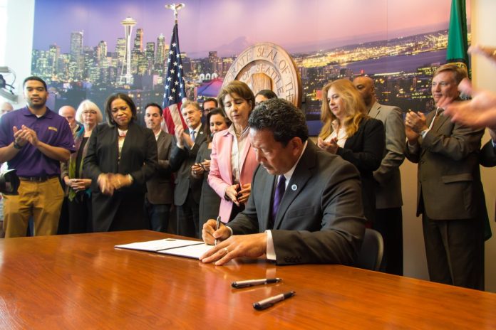Bruce Harrell sits at a desk signing some orders at City Hall with a dozen supporters looking on.