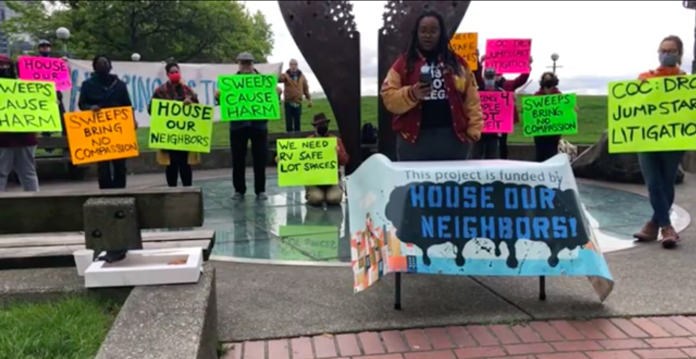 A photograph showing participants holding colorful signs at a press event. The signs carry messages such as 