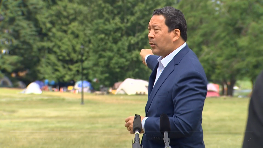 Harrell wears a blazer and gestures toward tents in the background.