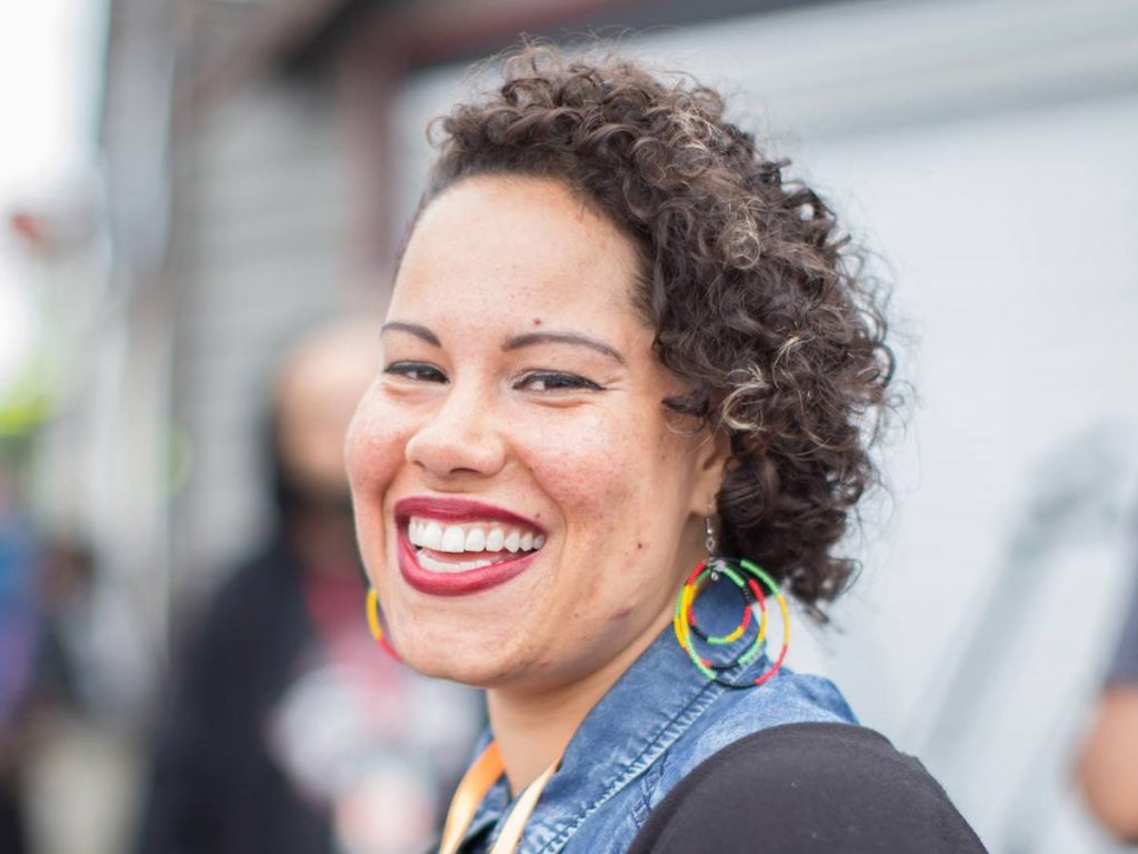 Nikkita is a Black woman. She wears rainbow hoop earrings. her curly hair to the side, and a big smile.