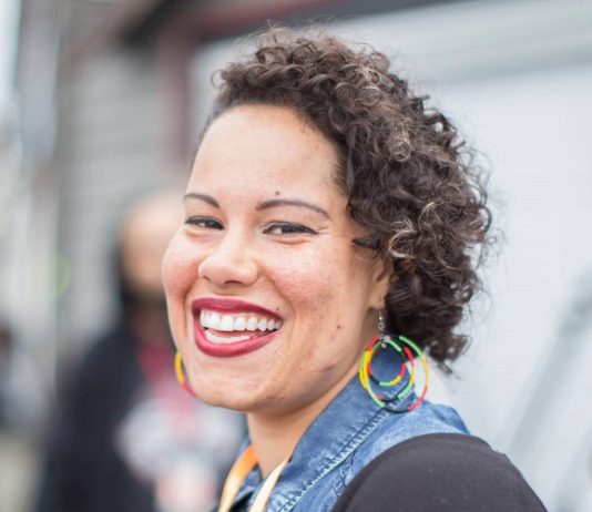 Nikkita Oliver Is Our Urbanist Meetup Guest on September 14th Nikkita is a Black woman. She wears rainbow hoop earrings. her curly hair to the side, and a big smile.