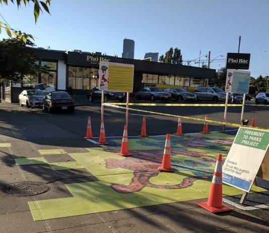 Seattle’s Neighborhood Recovery Fund Open for Proposals A photograph of a parking lot that has been painted with a brightly colored mural. Traffic cones mark the area surrounding part of the mural, and an A frame sign reads "pavement to parks."