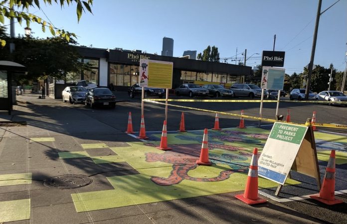 A photograph of a parking lot that has been painted with a brightly colored mural. Traffic cones mark the area surrounding part of the mural, and an A frame sign reads 