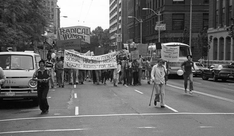 A black and white photo shows people marching through down town Seattle carrying signs. One large sign reads "Washington Coalition for Sexual Minority Rights, Equal Rights for All." Another sign reads, "Radical Women."