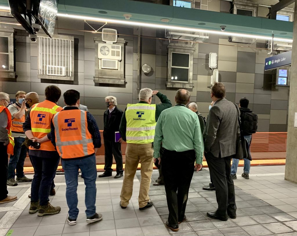 A group of men wearing colorful Sound Transit vests and suits stand at the station platform and look at the metal wall with architectural fragment inspired sculptures. 