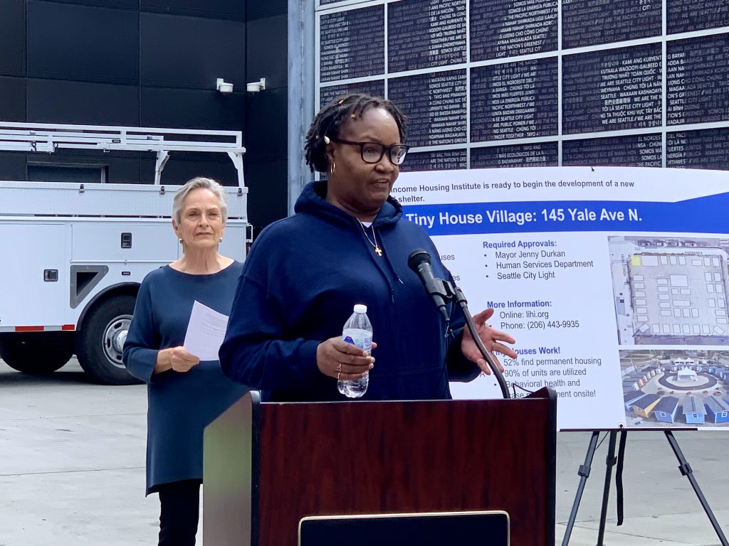 A woman wearing a dark blue hoodie speaks at podium. A sign describing the tiny house village is behind her. 