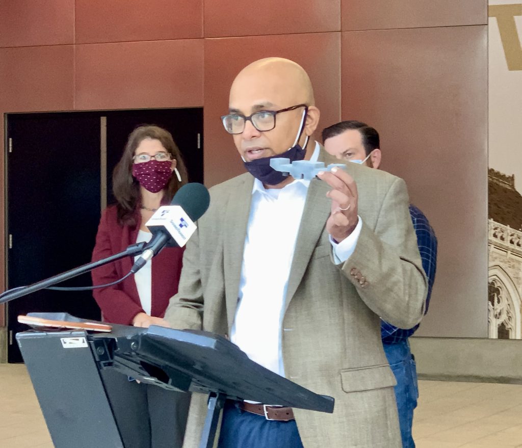 A brown skinned man wearing glasses and a face mask stands at a podium holding a piece of concrete slab. 