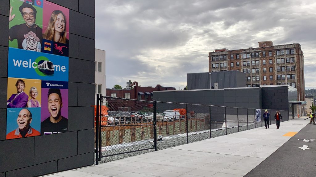 A photo of the two station buildings separated by an empty lot. A welcome sign with colorful photos is posted on the closer building. 