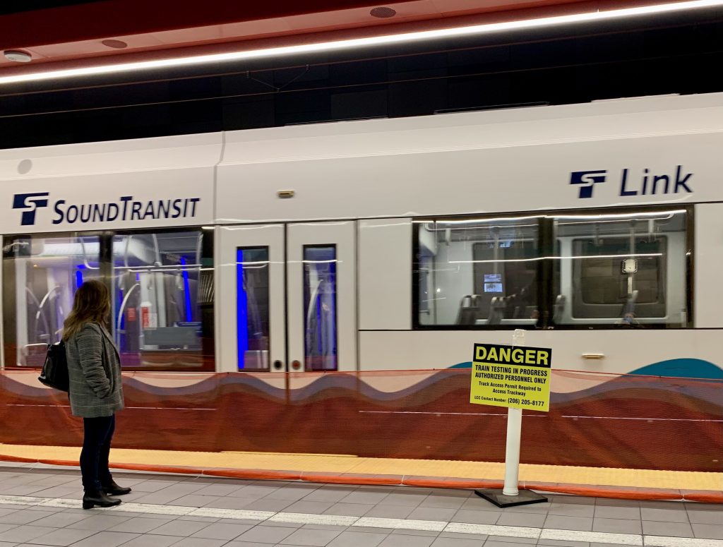 A woman stands in front of an empty Link light rail train. A yellow danger sign warns people that testing is in progress.
