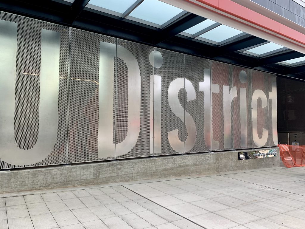  A photo shows an exterior metal wall with U District engraved in large letters. 