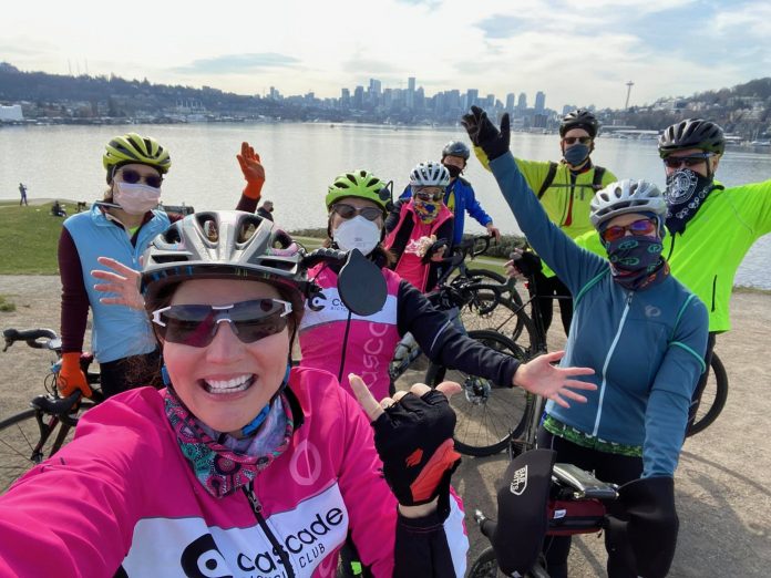 A group of cyclists wearing colorful athletic clothes poses near their bicycles in front of Lake Union in Seattle.