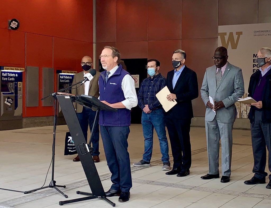 A photo of a White man standing at a podium wearing a blue Sound transit vest. A group of men stand behind him at the U District station entrance. 