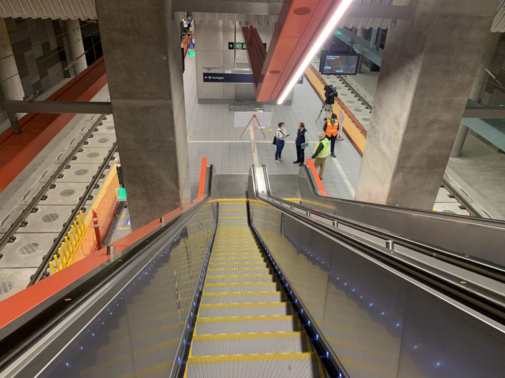 A photo looks down at the station platform from a shorter escalator. 