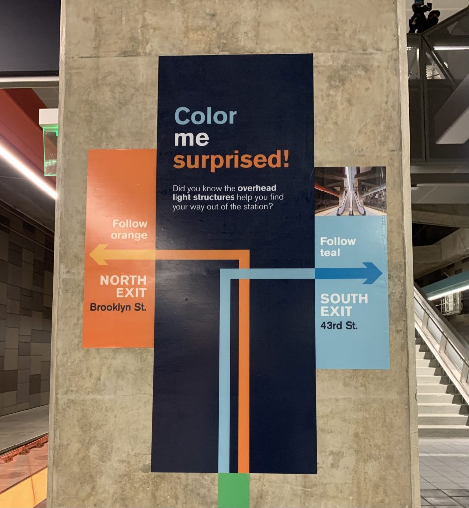 A wayfinding sign shows riders to follow the orange arrows and lights to the north exit at Brooklyn Street and the teal arrows and lights to the South exit at 43rd St. 