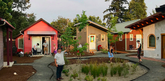 Op-Ed: How Mobile Villages Could Tackle Seattle’s Homelessness Emergency Tiny houses are oriented around a central green space where four people are standing. A couple is standing on the doorstep of a home in the background.