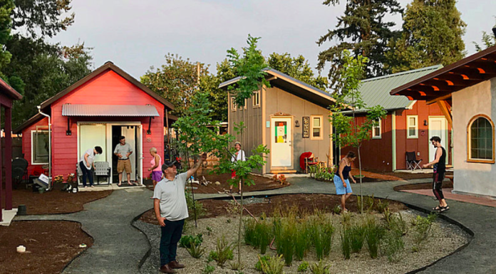 Op-Ed: How Mobile Villages Could Tackle Seattle’s Homelessness Emergency Tiny houses are oriented around a central green space where four people are standing. A couple is standing on the doorstep of a home in the background.
