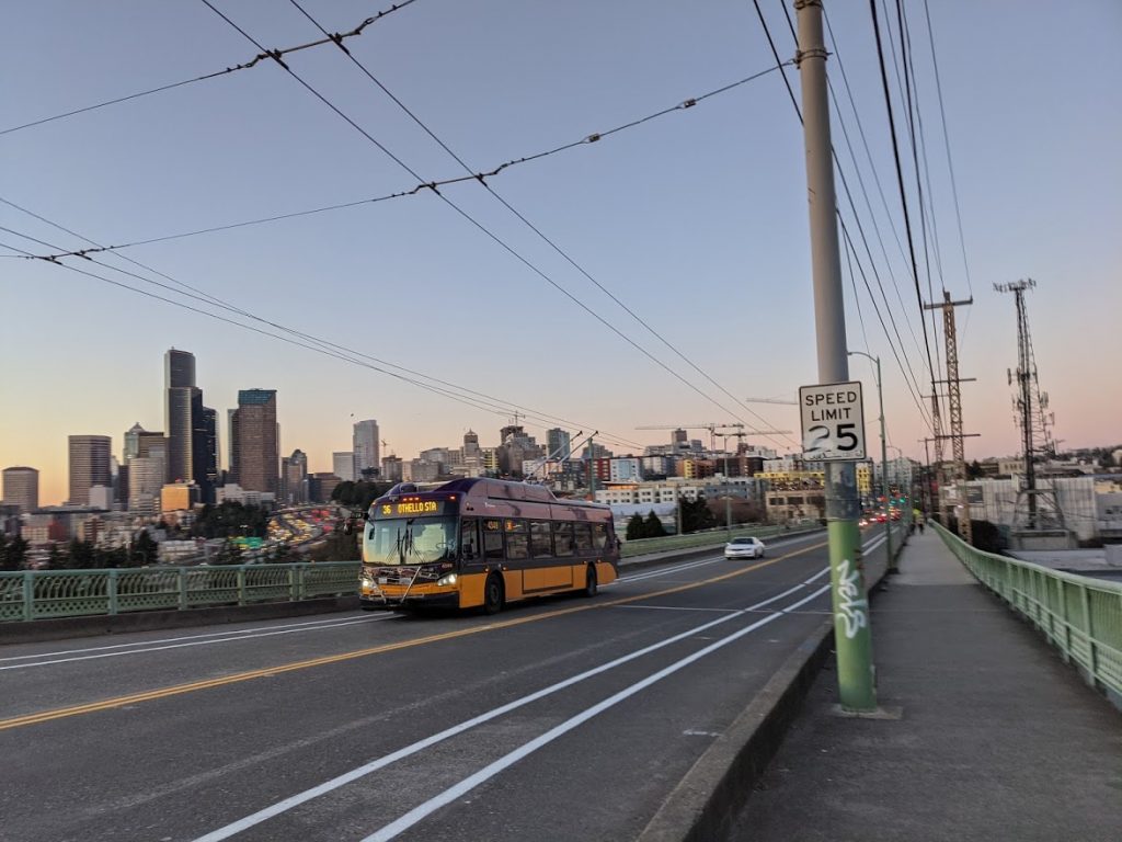 A photo shows a bike lane under construction on 12th Avenue at Jose Rizal Bridge. A King County Metro bus is driving on the bridge and the skyline of Downtown Seattle appears in the background. 