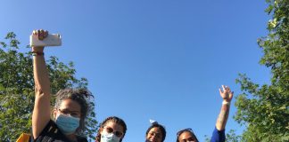 Five women wearing face masks raise their arms triumphantly in the air. They are standing outside with a blue sky behind them.
