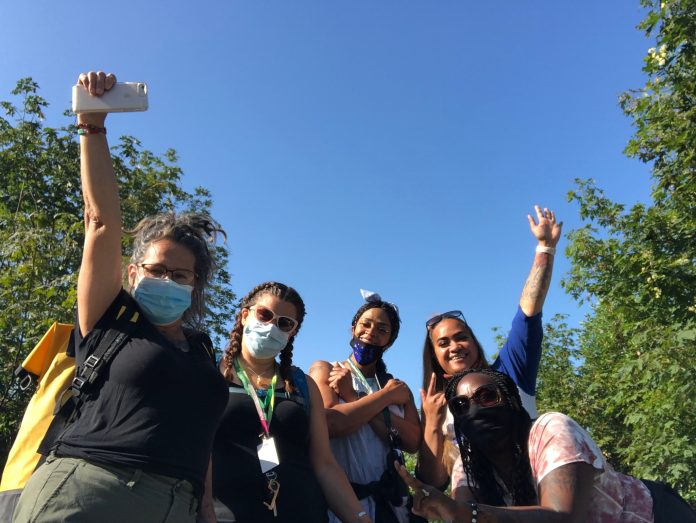 Five women wearing face masks raise their arms triumphantly in the air. They are standing outside with a blue sky behind them.