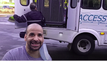 A bearded man in a tee shirt sits in front of a paratransit bus. 