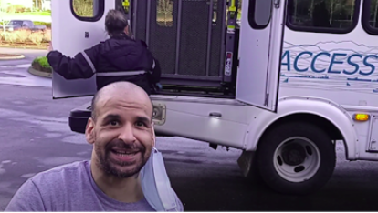 A bearded man in a tee shirt sits in front of a paratransit bus.