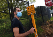 Report Calls for Radical Inclusion of Disabled Nondrivers in Transportation Planning Image description: A brown-skinned man standing at a highway offramp holding a white cane, next to a small yellow traffic sign with an icon of a pedestrian crossing and a bicycle reading “Use Caution When Crossing.” Behind that is a large red and white sign reading “Do Not Enter” and below that a small white sign with black text reading “Pedestrians Hitchhikers Bicycles Prohibited.”