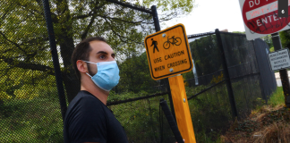 Image description: A brown-skinned man standing at a highway offramp holding a white cane, next to a small yellow traffic sign with an icon of a pedestrian crossing and a bicycle reading “Use Caution When Crossing.” Behind that is a large red and white sign reading “Do Not Enter” and below that a small white sign with black text reading “Pedestrians Hitchhikers Bicycles Prohibited.”