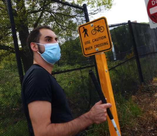 Report Calls for Radical Inclusion of Disabled Nondrivers in Transportation Planning Image description: A brown-skinned man standing at a highway offramp holding a white cane, next to a small yellow traffic sign with an icon of a pedestrian crossing and a bicycle reading “Use Caution When Crossing.” Behind that is a large red and white sign reading “Do Not Enter” and below that a small white sign with black text reading “Pedestrians Hitchhikers Bicycles Prohibited.”
