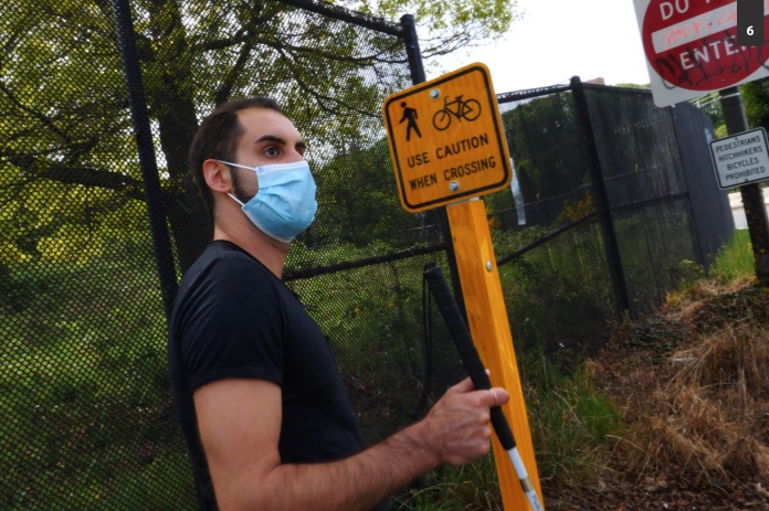 Image description: A brown-skinned man standing at a highway offramp holding a white cane, next to a small yellow traffic sign with an icon of a pedestrian crossing and a bicycle reading “Use Caution When Crossing.” Behind that is a large red and white sign reading “Do Not Enter” and below that a small white sign with black text reading “Pedestrians Hitchhikers Bicycles Prohibited.”