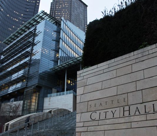 Lack of Clarity in Seattle City Code Harms Renters A photo of the Seattle City Hall with tall glass buildings illuminated at dusk in the background.