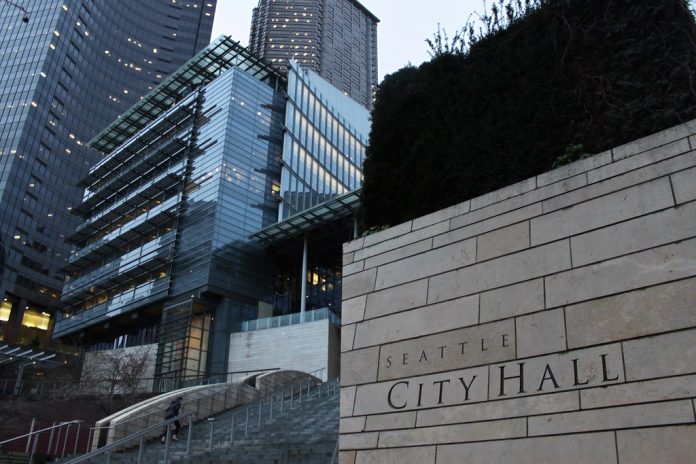 A photo of the Seattle City Hall with tall glass buildings illuminated at dusk in the background.