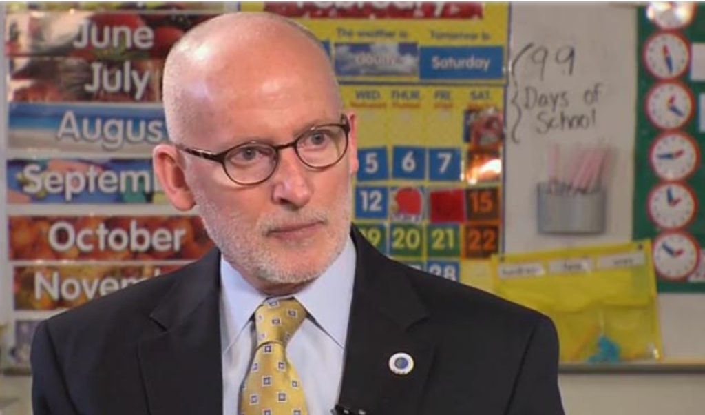 Tim Burgess is a bald white man with glasses dressed in a dark suit and yellow tie.