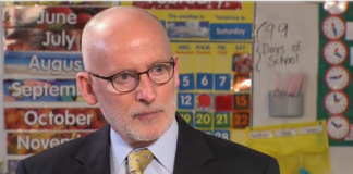 Tim Burgess is a bald white man with glasses dressed in a dark suit and yellow tie.