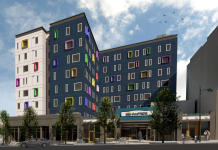 Pride Place Senior Housing Breaks Ground in Capitol Hill A rendering of a "L" shaped seven story apartment building. Three quarters of the exterior of the building is dark grey, while one quarter is white. The windows are surrounded by frames in many different vibrant colors.