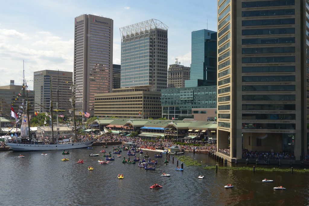 A photo of Harborplace shows a tall ship docked in the harbor surrounding by small colorful boats. Tall modern buildings line the waterfront. 