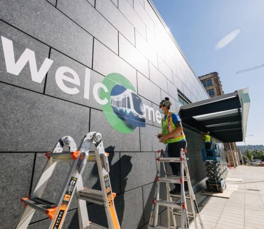 Celebrate Northgate Link Light Rail Expansion with Opening Day Events A photo of a worker standing on a ladder and stenciling welcome on to the exterior wall of the U District light rail station.