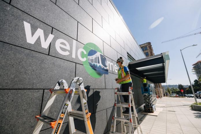 A photo of a worker standing on a ladder and stenciling welcome on to the exterior wall of the U District light rail station.