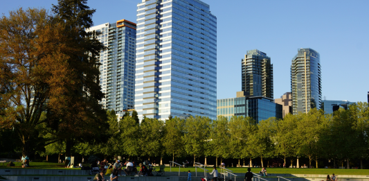 A photo of amphitheater style seating in a park with people sitting in the grass and tall modern buildings in the backdrop.