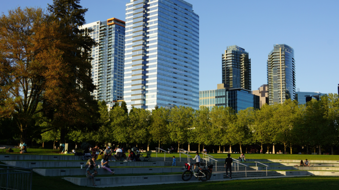 A photo of amphitheater style seating in a park with people sitting in the grass and tall modern buildings in the backdrop.
